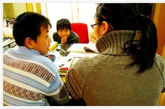 一名戴眼镜的女义工侧身为一个男孩讲题，另一个男孩在桌子另一头看书